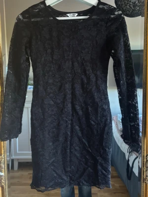 Svart spetsklänning med lång ärm - Snygg svart spetsklänning från Cubus med långärmad design och rund halsringning. Klänningen har ett elegant spetsmönster över hela plagget och är knälång. Perfekt för dig som gillar en klassisk och stilren look.