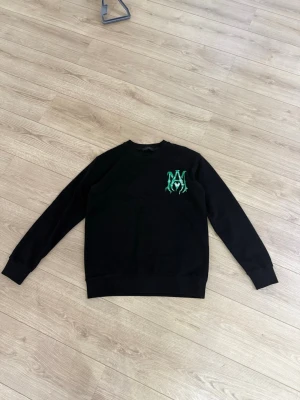 Svart sweatshirt från Amiri - Svart sweatshirt från Amiri med grönt tryck på bröstet och stor AMIRI-logga i grönt på ryggen. Tröjan har rund hals, långa ärmar och ribbade muddar. Perfekt för en streetwear-look.