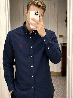 Två skjortor från Polo Ralph Lauren - Två klassiska skjortor från Polo Ralph Lauren, en i mörkblått med röd broderad logga och en i ljusgrått med marinblå logga. Båda har button-down krage och är tillverkade i mjuk bomull. Perfekta för en stilren look.                                  Priset är förhandlingsbart vid snabb överlåtelse. 
