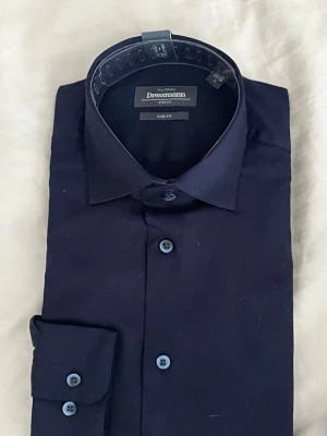  mörkblå slim fit skjorta Dressmann - Säljer en använd mörkblå slim fit skjorta från Dressmann med stretchmaterial. Skjortan har klassisk krage, blå knappar och lång ärm. Perfekt för dig som gillar en stilren look och bekväm passform. Den är Öpnna från sin paketering och har använts men är helt ny.Pris kan diskuteras. MVH stentorparchive⚓️