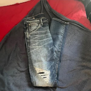 Jack & Jones ”Slim Glenn” - Säljer ett par blå skinny jeans med slitningar vid knät. Skick 9/10 och är i storlek 29/34. Jeansen har klassisk femficksdesign, låg midja och är tillverkade i denim. Perfekta för en avslappnad och trendig look.