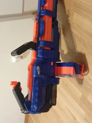 Nerf Titan CS-50 leksaksvapen - Säljer en Nerf Titan CS-50, ett stort leksaksvapen i blått och orange plast. Den har en rund trumma för skumdart och ett svart bärhandtag. Perfekt för intensiva Nerf-battles och har en cool design med detaljer i svart och transparent plast.