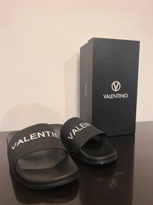 Svarta slides Valen - Säljer ett par svarta slides från Valenmed bred rem och vit logotyp på ovansidan. Sandalerna har en klassisk och stilren design, tillverkade i syntetmaterial med bekväm fotbädd. Perfekta för en avslappnad och trendig look. Passar även 45, Finn skador se bilder. Färg saknas.
