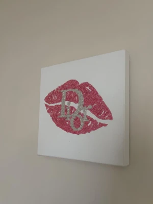 diamant Tavla med rosa läppar och glitter HANDGJORT - En handgjord tavla med motiv av rosa läppavtryck och glittriga bokstäver på vit canvas. Perfekt som dekoration i sovrum eller vardagsrum. Motivet är gjort med rosa och silverfärgade strasstenar för extra bling.
