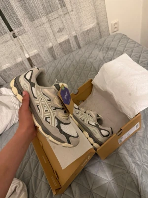 Asics gel nyc cream/oyster grey  43,5 storlek - Säljer ett par snygga ASICS sneakers i grått och vitt med mesh och syntetmaterial. Skorna har klassisk låg profil och coola detaljer på sidorna. Perfekt för dig som gillar sportig stil och vill ha bekväma skor med modern design.
