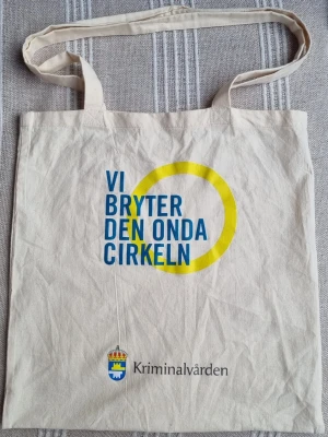 Tygkasse Kriminalvården med tryck - Beige tygkasse i bomull med blå och gul text och cirkel på framsidan. Trycket lyder 'Vi bryter den onda cirkeln' och har Kriminalvårdens logga längst ner.