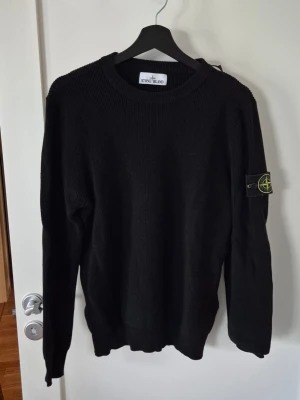 Svart stickad tröja Stone Island M - Säljer en svart stickad tröja från Stone Island med klassisk rund hals och ribbstickade muddar. Tröjan har den ikoniska Stone Island-loggan på vänster ärm. Perfekt för dig som gillar streetwear och stilrena plagg.