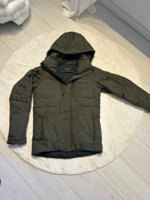 Olivgrön dunjacka Peak Performance - Snygg olivgrön dunjacka från Peak Performance med huva och flera dragkedjeförsedda fickor. Jackan har quiltad design och är perfekt för kalla vinterdagar. Klassisk logga broderad på bröstet och hög krage för extra värme.