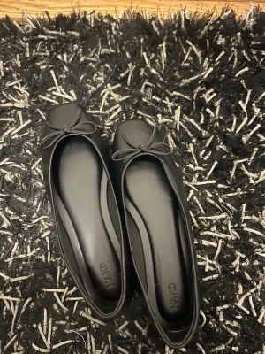 Svarta ballerinas från NA-KD - Stilrena svarta ballerinas från NA-KD med rund tå och dekorativ rosett framtill. Skorna har en klassisk form och är tillverkade i ett slätt syntetmaterial som ger en elegant look. Perfekta för dig som gillar enkel och tidlös stil.