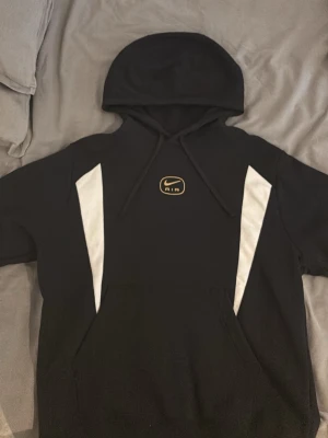Svart Nike Air hoodie med vita detaljer - Snygg svart hoodie från Nike Air med vita paneler längs sidorna och klassisk Nike Air-logga i guld på bröstet. Hoodien har huva med dragsko och stor magficka framtill. Perfekt för en sportig och avslappnad stil.