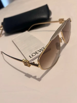 Guldiga aviator-solglasögon från Loewe - Snygga aviator-solglasögon från Loewe med guldfärgad metallbåge och bruna tonade glas. Glasögonen har unika detaljer vid skalmarna och en elegant design som sticker ut. Perfekta för dig som vill ha en lyxig accessoar till sommaren.Kvitto och all tillbehör medföljer,dem är mycket snyggare på -kan skicka bilder vid intresse.Nästan som nya.Kom med bud 