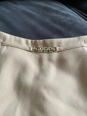 Beige kortkjol med kedjedetalj - Stilren beige kortkjol med snygg kedjedetalj framtill i midjan. Kjolen har en rak passform och är tillverkad i ett strukturerat tyg som ger en exklusiv känsla. Perfekt att matcha med en enkel topp eller stickad tröja.