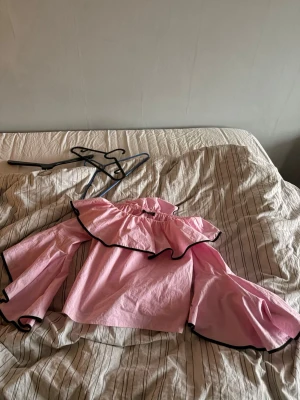 Rosa volangblus med svarta kanter - Trendig rosa blus med stora volanger runt axlarna och vida ärmar. Svarta kontrastkanter ger en snygg detalj. Off shoulder-modell med resår upptill för en bekväm passform. Perfekt för dig som vill sticka ut med en unik och lekfull stil.