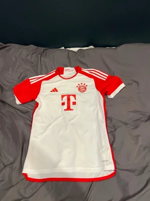 Bayern München matchtröja Adidas - Säljer en vit och röd matchtröja från FC Bayern München med Adidas-logga och klubbmärke på bröstet. Tröjan har korta ärmar, rund hals och klassiska Adidas-ränder på axlarna. Tillverkad i lätt och ventilerande material, perfekt för fotbollsträning eller match.