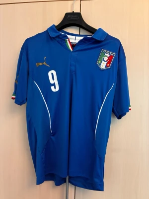 Italien Balotelli matchtröja Puma - Blå officiell Italien fotbollströja från Puma med nummer 9 och Balotelli på ryggen. Tröjan har Italiens landslagsemblem på bröstet, guldfärgad Puma-logga och detaljer i Italiens flaggfärger vid krage och ärmslut. Tillverkad i lätt och ventilerande material.