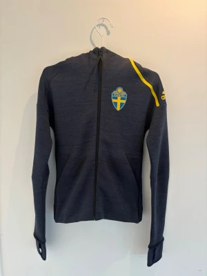 Blå adidas hoodie Sverige landslag - Mörkblå hoodie med dragkedja från adidas, prydd med Sveriges landslagsemblem på bröstet och gula detaljer på axeln samt adidas-logga på ärmen. Hoodien har fickor framtill och en skön huva. (Unisex) Aldrig använd men prislapp borttagen. Perfekt för dig som vill heja på Sverige i VM i sommar! 