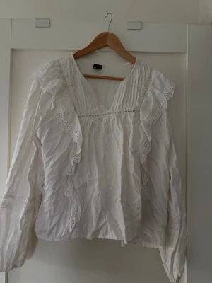 Vit blus med volanger från Gina Tricot - Säljer en vit blus från Gina Tricot i storlek XL. Blusen har långa ärmar, v-ringning och dekorativa volanger med spetsdetaljer längs fram och axlar. Materialet är lätt och luftigt, perfekt för att skapa en romantisk look.