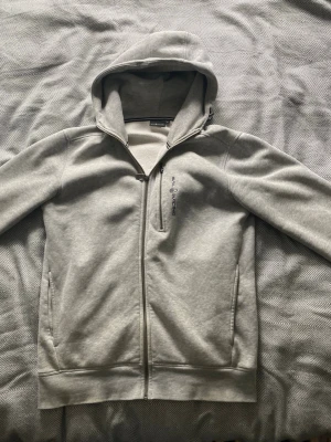 Ljusgrå hoodie från Sail Racing - Snygg ljusgrå hoodie från Sail Racing med dragkedja hela vägen, broderad logga på bröstet och praktisk bröstficka med dragkedja. Mjuk insida och klassisk huva gör den perfekt för chill dagar. Materialet känns som bomullsblandning.