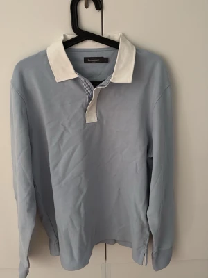 Ljusblå långärmad skjorta Dressmann - Ljusblå långärmad skjorta från Dressmann med vit krage och knappar. Skjortan har en klassisk design och är tillverkad i mjukt bomullsmaterial. Perfekt för dig som gillar stilrena plagg med en twist på kragen.