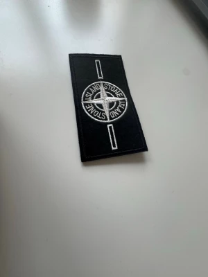 Stone island badge - white - Säljer en svart patch från Stone Island med broderad vit kompasslogga och vita detaljer. Perfekt att fästa på jackor, tröjor eller väskor för att ge din outfit en exklusiv touch. Materialet är textil och patchen har rektangulär form.