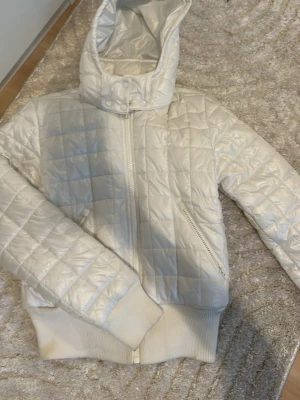 Vit quiltad pufferjacka från Adidas - Snygg vit pufferjacka från Adidas med quiltad design och huva. Jackan har dragkedja framtill, ribbade muddar och två sidofickor med dragkedja. Perfekt för kyliga dagar och ger en sportig vibe.