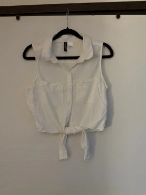 Vit ärmlös knytblus  - Säljer en vit ärmlös blus från Divided med krage, knappar framtill och två bröstfickor. Toppen har en snygg knytning nedtill som ger en avslappnad och trendig look. Perfekt till sommaren och lätt att matcha med jeans eller kjol. ( tänk på jag har hund i hemmet) 