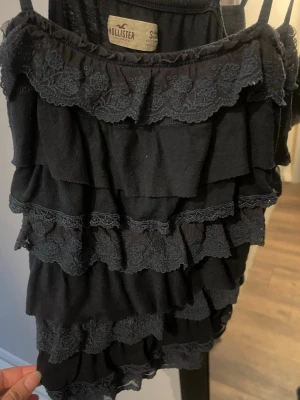 Mörkblå topp - Hollister - Mörkblå linnetopp från Hollister med tunna axelband och flera lager av volanger med spetsdetaljer. Perfekt för dig som gillar vintage toppar. Materialet är mjukt och lätt, vilket gör toppen bekväm att bära.