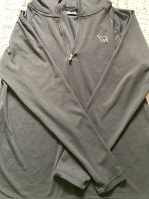Svart hoodie med dragkedja från WRP - Svart hoodie från WRP med halv dragkedja framtill och diskret logga på bröstet. Tillverkad i mjukt, stretchigt material och har en klassisk huva. Perfekt för en avslappnad och sportig stil.