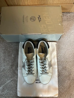 Golden Goose - Golden Goose sneakers i vitt skinn med grå mockastjärna på sidan och grå snörning. Skon har svart detalj på hälen och klassisk platt sula. Medföljer originalkartong och dustbag. Perfekta för dig som gillar en clean men ändå unik look. Bara höra av dig vid frågor.
