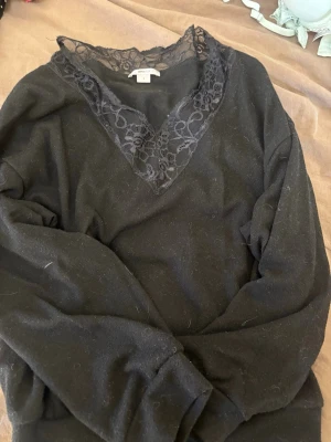 Svart tröja med spets från Gina Tricot - Snygg svart långärmad tröja från Gina Tricot med v-ringning och dekorativ spetsdetalj runt halsen. Tröjan har en mjuk känsla och passar perfekt till ett par jeans eller kjol. Spetsen ger en extra touch till looken.