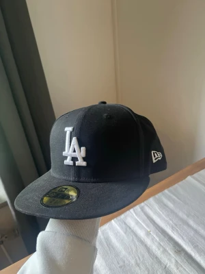 Svart LA-keps från New Era - Säljer en svart New Era 59FIFTY keps med LA Dodgers-logga broderad i vitt framtill och MLB-logga bak. Klassisk flat brim och strukturerad passform. Perfekt för dig som gillar streetwear och sportig stil.