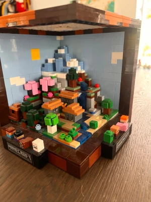 Minecraft LEGO diorama - Säljer ett detaljerat Minecraft LEGO diorama med berg, träd, vattenfall och blommor i olika färger. Modellen är byggd i en öppen kub med bruna, blå, gröna och rosa klossar. Perfekt för dig som gillar Minecraft och LEGO.