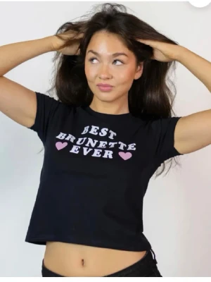 Svart croppad t-shirt med tryck - Svart croppad t-shirt med vit text 'Best Brunette Ever' och rosa hjärtan på bröstet. T-shirten har korta ärmar och en tight passform. Perfekt för dig som vill ha en statement-tee med attityd.