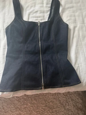 Mörkblå denimtopp från Zara - Säljer en snygg mörkblå denimtopp från Zara med markerade sömmar och dragkedja framtill. Toppen har breda axelband och en figurnära passform.