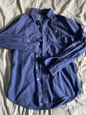 Marinblå skjorta från Ralph Lauren - Klassisk marinblå skjorta från Ralph Lauren med den ikoniska gröna broderade loggan på bröstet. Skjortan har lång ärm, knappar framtill och button-down krage. Tillverkad i mjuk bomull för en bekväm känsla och snygg passform.