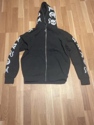Svart zip hoodie från Black Squad - Svart hoodie med dragkedja från Black Squad i relaxed fit. Hoodien har vita tryck längs båda ärmarna och på huvan, samt två fickor framtill. Tillverkad i mjukt material, perfekt för en chill och streetwear-inspirerad look.