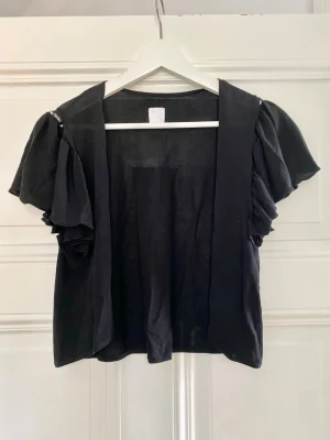 Söt bolero med fina detaljer - Söt svart bolero/ öppen blus med fina detaljer på ärmarna (se bild nr2). Osäker på klädmärkets namn då loggan ej innehåller hela namnet (se sista bilden), men vet att den är från ett danskt märke och köpt för ca 1000-1500kr. 