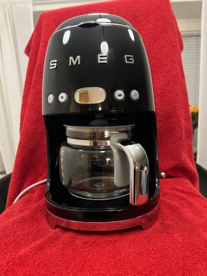 "Smeg" Kaffebryggare DCF02BLEU Svart - Stilren svart kaffebryggare från Smeg med retrovibbar. Maskinen har rundad form, tydliga knappar på framsidan och en glaskanna med kromat handtag. Vattennivåindikator finns på sidan och detaljer i metall ger en lyxig känsla. Perfekt för köket om du gillar snygg design.     *** Knappt använt jättefint kondition. Säljer på grund av jag fick en ny nya Moccamaster.