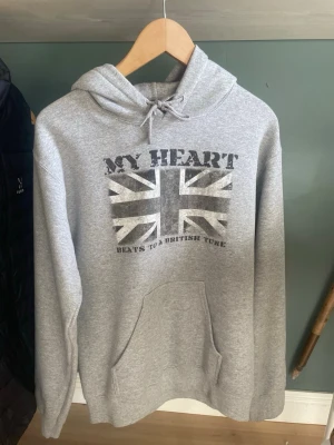 Drakenberg MY HEART hoodie - Snygg grå hoodie med tryck av Union Jack och texten 'MY HEART BEATS TO A BRITISH TUNE' på bröstet. Klassisk känguruficka framtill och justerbar huva med dragsko. Perfekt för dig som gillar streetwear och brittisk stil. Drakenberg. Helt ny, säljer pågrund av att den var för liten och orkar inte skicka tillbaka.