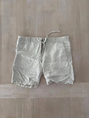 Beiga linneshorts med snörning - Säljer ett par ljusbeige shorts i linne med snörning i midjan och fickor både fram och bak. Shortsen har en avslappnad passform och bälteshällor för extra stilpoäng. Perfekta för varma dagar och enkel att matcha med olika toppar.