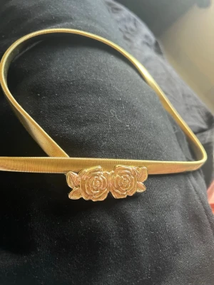 Guldigt bälte med rosdetalj - Snyggt och smalt bälte i guld med en dekorativ spänne i form av två rosor. Perfekt accessoar för att lyfta vilken outfit som helst med en elegant touch. Metallisk finish och flexibel design.
