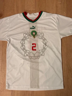 Marocko fotbollströja Puma vit - Säljer en vit Marocko fotbollströja från Puma med röd och grön krage, nummer 2 i rött på bröstet och Marockos fotbollsförbunds emblem. Tröjan har ett unikt geometriskt mönster på framsidan och är tillverkad i lätt, andasvänligt material. XL men passar även för L