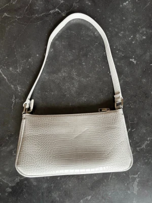 Vit axelremsväska med krokopräglat mönster - Snygg vit handväska i avlång modell med axelrem och krokopräglat mönster. Väskan har silverfärgade detaljer och dragkedja upptill. Perfekt accessoar för att lyfta din outfit med en trendig touch. Vet inte vart den är ifrån. Det är min tjejs. 