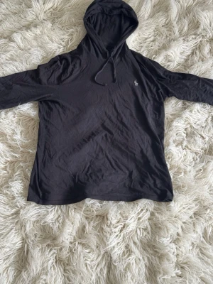 Svart hoodie från Ralph Lauren - Säljer en tunn svart hoodie från Ralph Lauren med klassisk huva och dragsnören. Perfekt för en avslappnad och stilren look. Passar storlek M.