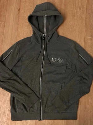Mörkgrå hoodie från Hugo Boss - Snygg mörkgrå hoodie från Hugo Boss med dragkedja framtill och logga på bröstet. Tröjan har huva med dragsko, ribbade muddar och diskreta zip-detaljer på ärmarna. Perfekt för en avslappnad och stilren look. Den är M men passar även för S 