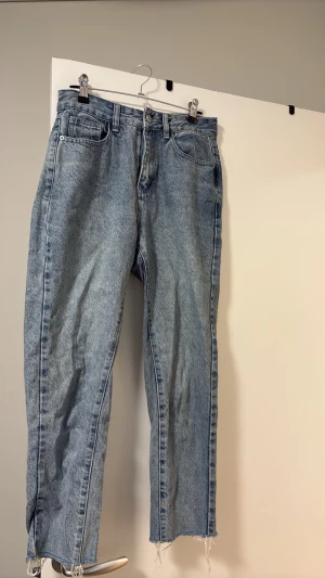 Ljusblå raka jeans med rå kant - Säljer ett par ljusblå jeans med rak passform och rå, fransig kant nedtill. Klassisk femficksmodell och hög midja. Perfekta för en avslappnad och trendig look. Materialet är denim i bomull.