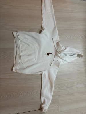 Vit hoodie från Polo Ralph Lauren - Säljer en vit hoodie från Polo Ralph Lauren med den klassiska broderade polospelaren på bröstet. Hoodien har huva med dragsko och en stor magficka framtill. Tillverkad i mjuk bomull, perfekt för en chill och stilren look.