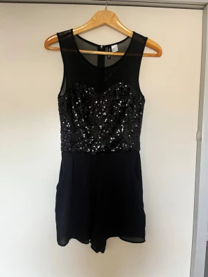 Svart paljettjumpsuit från H&M - Snygg svart jumpsuit från H&M med transparent mesh upptill och paljetter över hela överdelen. Kort modell med rund halsringning och dragkedja bak. Perfekt för dig som vill sticka ut lite extra. 