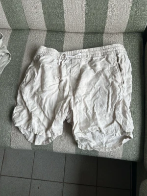 Ljusbeige linneshorts med resår - Säljer ett par ljusbeige shorts i linne med elastisk midja och sidofickor. De har en avslappnad passform och en bakficka. Perfekta för varma dagar och ger en chill vibe till din outfit.