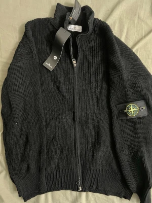 Svart stickad ziptröja Stone Island - Säljer en svart stickad ziptröja från Stone Island med hög krage och ribbade muddar. Tröjan har dragkedja framtill och den klassiska Stone Island-loggan på ärmen. Perfekt för dig som gillar streetwear och vill ha något stilrent och bekvämt.